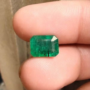 Puede incluir: Una esmeralda rectangular de color verde intenso. La gema tiene un corte facetado que refleja la luz. Se aprecian inclusiones internas. La esmeralda se sostiene entre los dedos, mostrando su color y claridad.
