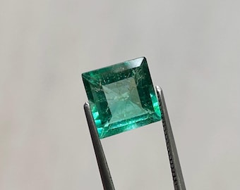 Puede incluir: Una esmeralda verde vibrante, de corte cuadrado, es sostenida por unas pinzas plateadas. La esmeralda tiene una superficie facetada, que refleja la luz y muestra su claridad. La gema es de un color verde intenso.