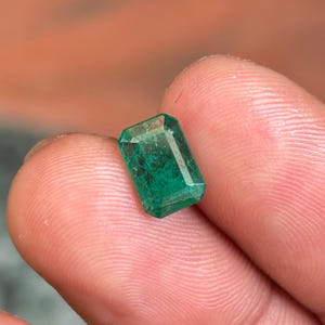 Könnte beinhalten: Ein rechteckiger Smaragd-Edelstein mit leuchtend grüner Farbe. Der Stein hat einen facettierten Schliff, der seine Klarheit und inneren Muster zeigt. Der Smaragd wird zwischen Fingern gehalten, was seine Größe und Form hervorhebt.