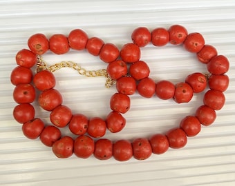 Puede incluir: Un collar largo hecho de cuentas de coral rojo brillante. Las cuentas son redondas y tienen una superficie rugosa y texturizada. El collar está enhebrado en una cadena de oro.