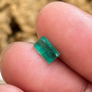May include: A rectangular, emerald-green gemstone with visible internal structures. The gem is held between two fingers, showcasing its clarity and color. The stone appears to be a raw, uncut emerald.