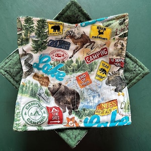 May include: A quilted bowl cozy with a camping theme. The fabric features a map design with bears, deer, and signs that say "Camping", "Bear in Area", and "Tent Sites". The cozy is white with green accents.
