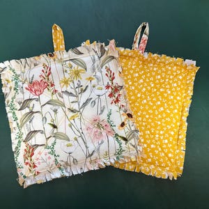 May include: Two square pot holders with ruffled edges. One features a floral pattern with pink, red, and yellow flowers on a white background. The other is yellow with a white floral pattern. Both have hanging loops.