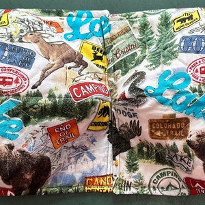 May include: A quilted fabric bowl cozy with a camping theme. The white fabric is patterned with images of deer, bears, trees, and camping signs. The word "Lake" is stitched in turquoise across the front. The cozy is designed to hold a bowl of hot food.