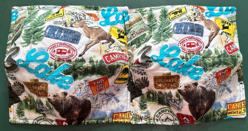 May include: A quilted fabric bowl cozy with a camping theme. The white fabric is patterned with images of deer, bears, trees, and camping signs. The word "Lake" is stitched in turquoise across the front. The cozy is designed to hold a bowl of hot food.