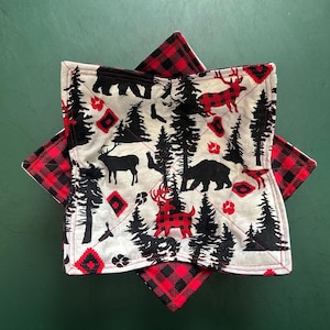 May include: Two square quilted bowl cozies. One features a black and red buffalo plaid pattern. The other has a white background with black trees, bears, deer, and red accents. The cozies are designed to hold hot bowls.