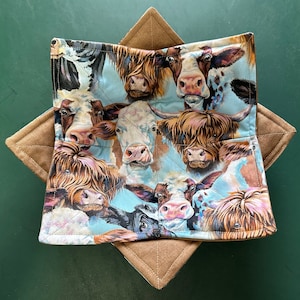 May include: A blue and white fabric pot holder with a patchwork design featuring various breeds of cows. The cows are depicted in a realistic style with brown, white, and black markings.