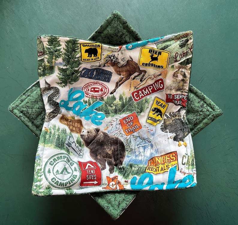 May include: A quilted bowl cozy with a camping theme. The fabric features a map design with bears, deer, and signs that say "Camping", "Bear in Area", and "Tent Sites". The cozy is white with green accents.