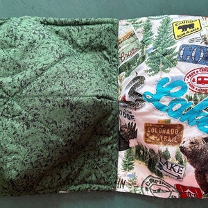 May include: A quilted bowl cozy with two sides. One side is green with a leaf pattern, the other features a camping theme with images of a bear, deer, and the word "Lake".