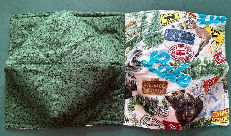 May include: A quilted bowl cozy with two sides. One side is green with a leaf pattern, the other features a camping theme with images of a bear, deer, and the word "Lake".