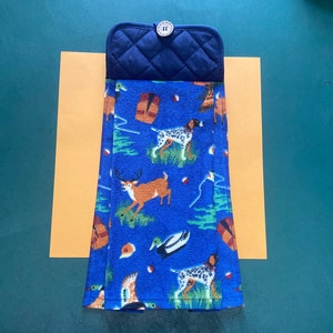 May include: A blue kitchen towel with a hunting theme. The towel features images of dogs, deer, ducks, and other wildlife. The towel is folded and has a dark blue quilted potholder attached to the top.