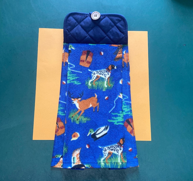 May include: A blue kitchen towel with a hunting theme. The towel features images of dogs, deer, ducks, and other wildlife. The towel is folded and has a dark blue quilted potholder attached to the top.