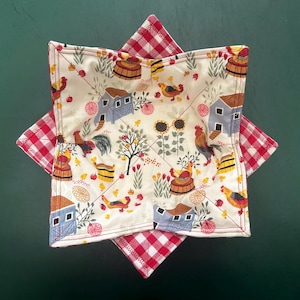 May include: A quilted bowl cozy with a farm-themed pattern. The cozy features a cream-colored fabric with illustrations of roosters, chickens, barns, and sunflowers. The edges are trimmed with red and white gingham fabric.