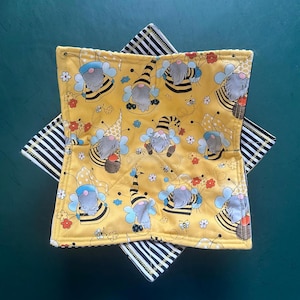 May include: A square fabric bowl cozy featuring a yellow background with a repeating pattern of gnomes dressed as bees with wings and striped hats, accented by small flowers and bees. The underside is black and white striped fabric with visible stitching.