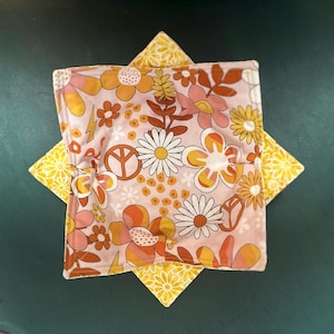 May include: A floral print bowl cozy with a pink background and a yellow starburst base. The cozy features a retro floral pattern with daisies, orange flowers, and a peace sign. The base has a yellow floral pattern.