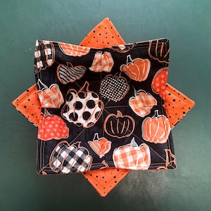 May include: A black and orange quilted bowl cozy with a pumpkin pattern. The cozy features various pumpkin designs in orange, white, and black, with a contrasting orange polka dot backing. The cozy is designed to hold a bowl.