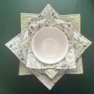 May include: A stack of white ceramic dishes and a bowl, resting on square, quilted placemats. The placemats feature a white background with a green leaf and pink berry pattern. The placemats are layered on top of each other.