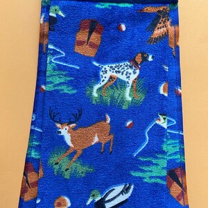 May include: Blue kitchen towel with a repeating pattern of dogs, deer, ducks, and other hunting-themed imagery. The towel is folded and has a blue quilted fabric backing with a button.
