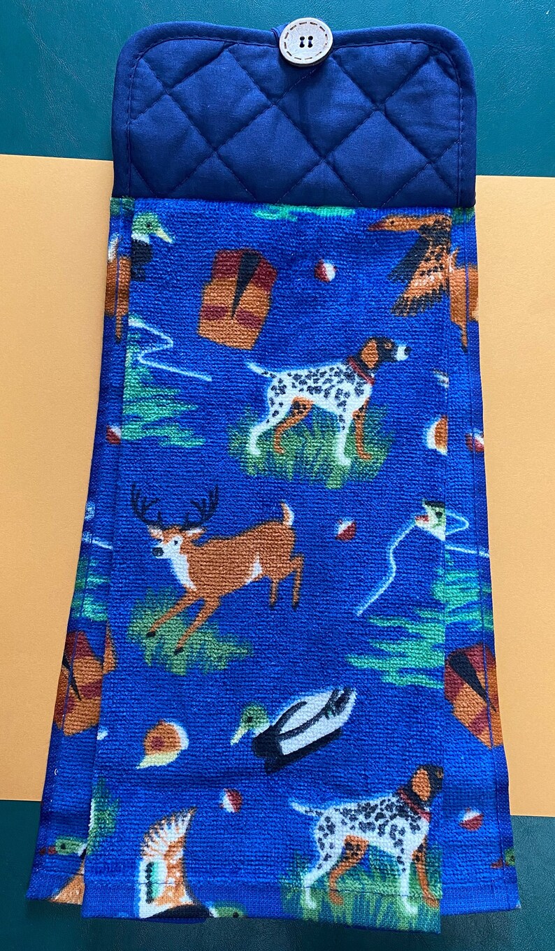 May include: Blue kitchen towel with a repeating pattern of dogs, deer, ducks, and other hunting-themed imagery. The towel is folded and has a blue quilted fabric backing with a button.
