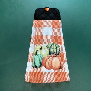 May include: A kitchen towel with a checkered orange and white pattern. The towel features a design of four pumpkins in shades of green, orange, and brown.