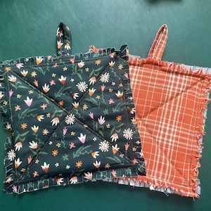 May include: Two square pot holders. One is dark green with a floral pattern and ruffled edges. The other is orange and white plaid with frayed edges. Both have a loop for hanging.