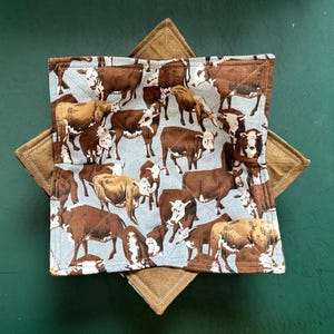 May include: A brown and tan quilted fabric bowl with a pattern of brown cows on a light blue background. The bowl is made of two layers of fabric, with the top layer being quilted and the bottom layer being a solid brown fabric.