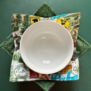 May include: A white ceramic bowl sits inside a green and white fabric bowl cozy. The cozy features a camping theme with images of trees, animals, and signs. The cozy is designed to protect hands from hot bowls.