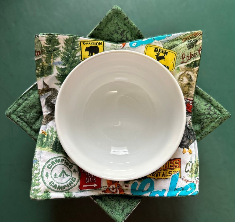 May include: A white ceramic bowl sits inside a green and white fabric bowl cozy. The cozy features a camping theme with images of trees, animals, and signs. The cozy is designed to protect hands from hot bowls.