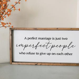 May include: Wooden framed sign with the text "A perfect marriage is just two imperfect people who refuse to give up on each other" in black script. The sign is on a light gray surface.