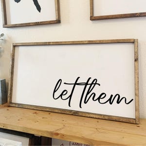 May include: A white wooden sign with a brown wood frame. The sign has black calligraphy that reads "let them".