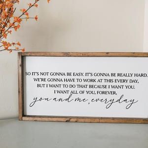 May include: A rectangular wooden framed sign with a white background and black text. The text reads, "So it's not gonna be easy...you and me, everyday." Decorative orange flowers are in a clear vase to the left.