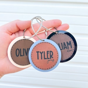 May include: Three round leather keychains with metal rings. The keychains are brown, blue, and black. The names "Olivia", "Tyler James", and "Liam Dean" are printed on the keychains.