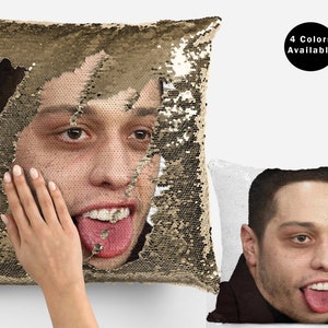 May include: A gold sequin pillow with a face of a man sticking out of it. The man is sticking his tongue out and has a mischievous look on his face. The pillow is reversible and the other side shows the same man's face with a white background. The text "4 Colors Available" is in a black circle in the upper right corner of the image.