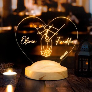 Pode incluir: Uma luz noturna de madeira em forma de coração com duas mãos se segurando. A luz tem os nomes "Olivia" e "Freddie" e a data "01/19/2023".