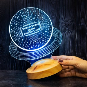 Könnte beinhalten: Eine blaue und weiße 3D-gedruckte Lampe mit einem Leiterplatten-Design. Der Lampenfuß ist aus Holz und hat eine runde Form. Die Lampe leuchtet und der Text "Electronics Electrical Engineer Jonathan Lovato" ist auf der Leiterplatte sichtbar.