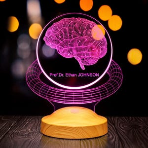 Könnte beinhalten: Eine rosa 3D-gedruckte Gehirndarstellung auf einem klaren Acrylsockel mit einem Holzsockel. Der Sockel trägt den Text "Prof. Dr. Ethan JOHNSON".