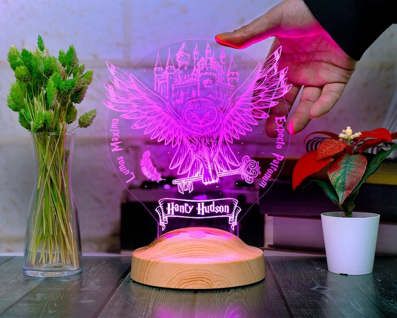 Peut inclure: Une veilleuse imprim&eacute;e en 3D rose et blanche avec un motif de hibou aux ailes d&eacute;ploy&eacute;es, un ch&acirc;teau et une cl&eacute;. La veilleuse a une base en bois et le texte "Lumos Maxima", "Expecto Patronum" et "Hony Hudson" dessus.