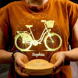 Könnte beinhalten: Ein gelbes, beleuchtetes Nachtlicht in Form eines Fahrrads mit einem Korb am Lenker. Das Licht hat einen Holzsockel und der Name "Sophia" ist auf der Vorderseite aufgedruckt.