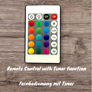 Peut inclure: Une t&eacute;l&eacute;commande blanche avec une vari&eacute;t&eacute; de boutons, y compris des options de couleur, une fonction flash et une minuterie. Le texte "OFF ON" et "FLASH" est visible sur la t&eacute;l&eacute;commande. Le texte "Remote Control with Timer function" et "Fernbedienung mit Timer" est visible sous la t&eacute;l&eacute;commande.