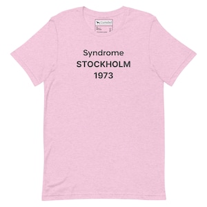 May include: A light pink short-sleeve t-shirt with a crew neckline. The center of the chest features dark gray text that reads Syndrome STOCKHOLM 1973. The interior neck label displays the Curtailed brand logo and size information.