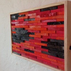 Op de afbeelding: Een rechthoekig houten wandkunstwerk met een mozaïek van kleurrijke houten blokken. De blokken zijn voornamelijk rood, oranje en zwart, gerangschikt in een horizontaal patroon binnen een licht houten frame. Het kunstwerk is gemonteerd op een getextureerde witte muur.