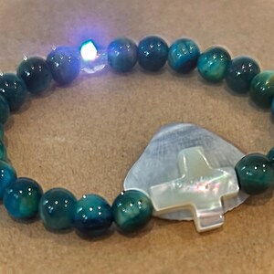 May include: A beaded bracelet with teal and dark green beads. The bracelet features a mother-of-pearl cross and a small, glowing light. The beads are round and have a glossy finish. The bracelet is resting on a light brown surface.