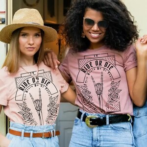 Pu&ograve; includere: Due donne indossano magliette rosa con un design grafico in bianco e nero che dice "Ride or Die" con una mano scheletrica che fa un segno rock and roll. La grafica &egrave; circondata da rose.