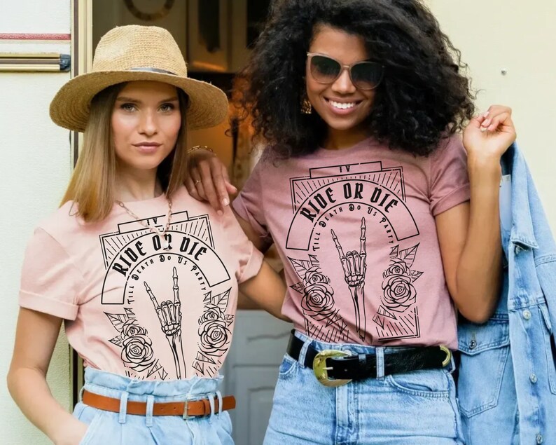Pu&ograve; includere: Due donne indossano magliette rosa con un design grafico in bianco e nero che dice "Ride or Die" con una mano scheletrica che fa un segno rock and roll. La grafica &egrave; circondata da rose.