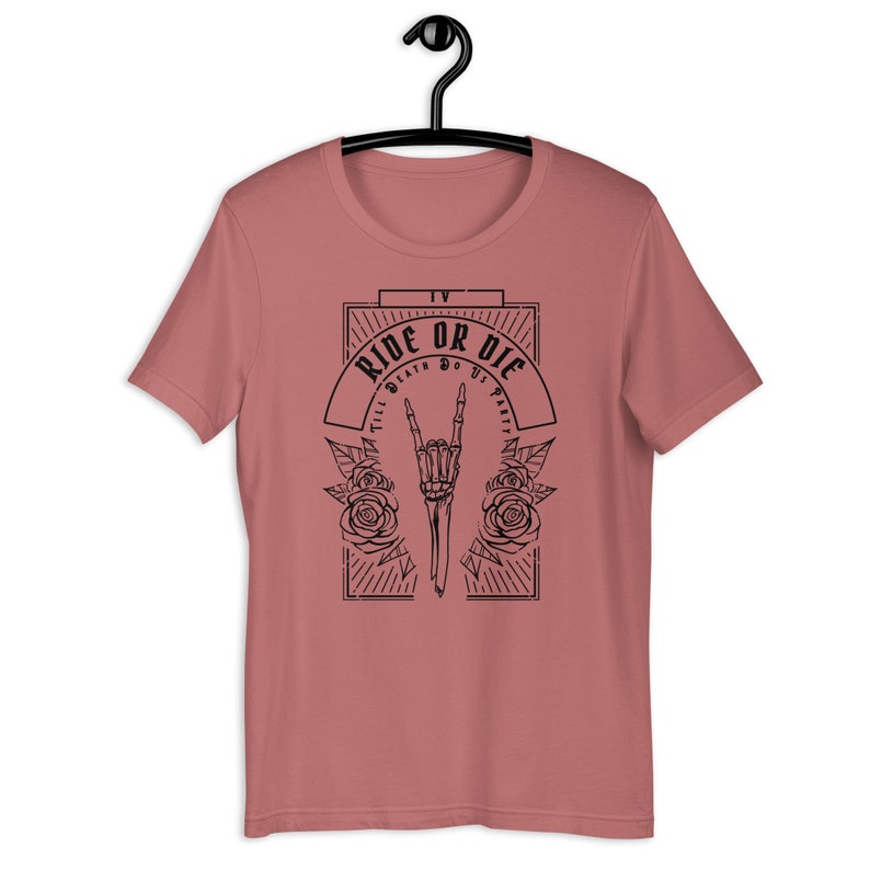 Pu&ograve; includere: Una t-shirt rosa con un design grafico nero che presenta una mano scheletrica che fa il segno del rock and roll. La grafica include anche il testo "Ride or Die" e "Till Death Do Us Party".