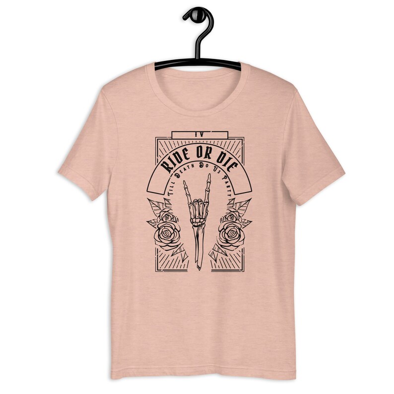Pu&ograve; includere: Una t-shirt rosa chiaro con un design grafico nero. La grafica presenta una mano scheletrica che fa un gesto rock and roll con rose e il testo "Ride or Die Till Death Do Us Part".