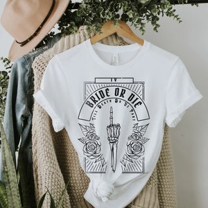 Pu&ograve; includere: Maglietta bianca con un design grafico nero che dice "Bride or Die" con una mano scheletrica che fa il dito medio e rose. Il testo sotto la grafica dice "Till Death Do Us Part."