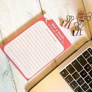 Op de afbeelding: Een roze notitieboekje met de tekst "Wait a minute... Let me just overthink this My notes and thoughts" bovenaan. Het notitieboekje ligt op een wit houten oppervlak met een zilveren laptop en roze paperclips.