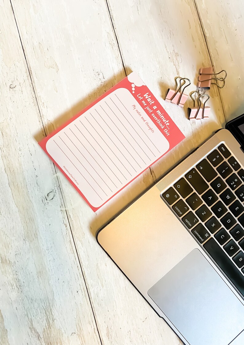 Op de afbeelding: Een roze notitieboekje met de tekst "Wait a minute... Let me just overthink this My notes and thoughts" bovenaan. Het notitieboekje ligt op een wit houten oppervlak met een zilveren laptop en roze paperclips.