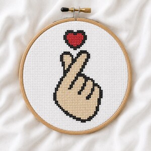 May include: A cross-stitch embroidery piece mounted in a circular wooden hoop. The design features a hand making a finger heart gesture, outlined in black with beige skin tones, positioned below a small, pixelated red heart on a white fabric.